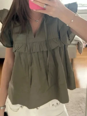 Olivgrön volangblus med kort ärm - Snygg olivgrön blus med volangdetaljer och korta ärmar. Blusen har v-ringning med knytband och rynkade partier framtill. Perfekt för en trendig och avslappnad look. Tillverkad i ett lätt och luftigt tyg.