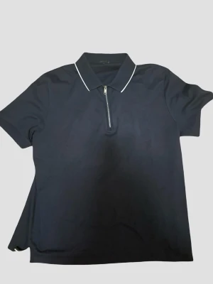 Marinblå golfpiké ifrån Boss - Riktigt snygg mörkblå golfpiké ifrån Boss med diskret logga och vit piping på kragen och halv dragkedja framtill. Kortärmad modell i mjukt material, perfekt för en clean och sportig look. XL men är liten i storleken (motsvarar snarare L i verkligheten).