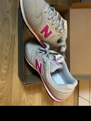 Gråa New Balance sneakers med rosa detaljer - Snygga sneakers från New Balance i grått med rosa N-logga på sidan och vita samt rosa detaljer. Skorna har klassisk låg profil, snörning och slitstark gummisula. Ovandelen är i syntetmaterial och mesh för bra andningsförmåga.