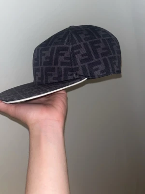 Fendi keps med FF-mönster - Säljer en grå keps från Fendi med det klassiska FF-logomönstret över hela kepsen. Kepsen har rak skärm och justerbar rem baktill för perfekt passform. Materialet är tyg med exklusiv känsla och insidan av skärmen är vit.