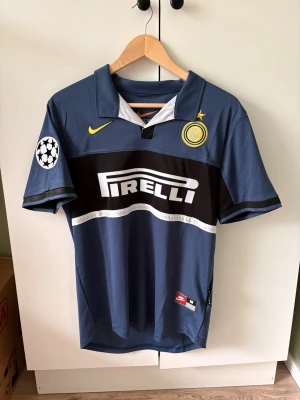 Inter Milan Baggio matchtröja Nike - Retro Inter Milan matchtröja i blått med svarta och vita detaljer, officiellt tryck med Baggio och nummer 10 på ryggen. Nike-logga och klubbmärke på bröstet, Pirelli som sponsor framtill och Champions League-märke på ärmen. Perfekt för fotbollsfans!