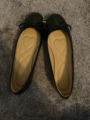 Svarta ballerinaskor med rosett - Stilrena svarta ballerinaskor med glansig finish och söt rosett framtill. Innersulan är ljusbeige med hjärtdetaljer för extra charm. Skorna har rund tå och platt sula, perfekta för en trendig och bekväm look.