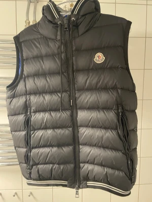 Svart dunväst från Moncler med huva - Snygg svart dunväst från Moncler med avtagbar huva och dragkedja framtill. Västen har två fickor med dragkedja och Moncler-logga på bröstet. Ribbstickade detaljer vid krage och nederkant ger en sportig look. Perfekt för lager-på-lager-stil.