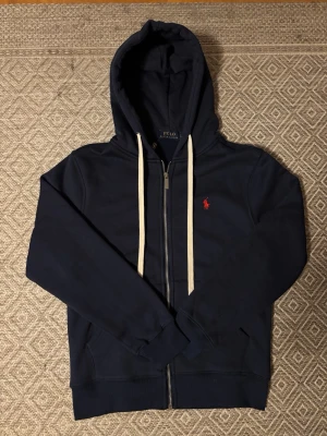 Mörkblå hoodie från Polo Ralph Lauren - Mörkblå hoodie med dragkedja från Polo Ralph Lauren. prillans ny med lappen kvar. Sitter dock inte kvar på hoodies som det syns i första bilden. Säljs för den var för liten för mig. Jag är 186.