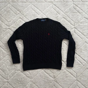 Polo Ralph Lauren Navy knitwear  - Kabelstickad svart tröja från Polo Ralph Lauren med rund halsringning och röd broderad logga på bröstet. Tröjan har långa ärmar och ribbade muddar vid ärmslut och nederkant. Klassisk och stilren design i mjukt material.