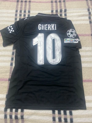 Svart Manchester City matchtröja Puma - Säljer en svart Manchester City matchtröja från Puma med silverfärgade detaljer och tryck. Tröjan har namn och nummer 'Cherki 10' på ryggen, Etihad Airways-logga på bröstet och Champions League-märke på ärmen. Tillverkad i funktionsmaterial, perfekt för fotboll eller träning.