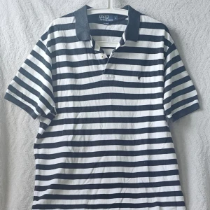 Randig pikétröja från Polo Ralph Lauren - Klassisk svartvit randig pikétröja från Polo Ralph Lauren med krage och knappar framtill. Tröjan har korta ärmar och den ikoniska broderade loggan på bröstet. Perfekt för en avslappnad men ändå stilren look.