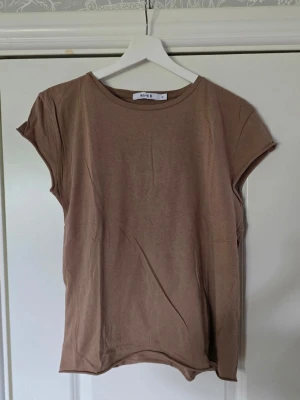 Beige t-shirt från NA-KD - En enkel och stilren beige t-shirt från NA-KD i 100% bomull. T-shirten har korta ärmar och rund halsringning. Perfekt basplagg till garderoben och lätt att matcha med olika outfits.
