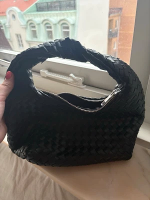 Svart flätad handväska - Cool svart handväska med flätad design och vriden axelrem. Väskan har en zip-stängning och är tillverkad i läder-look material. Perfekt för dig som vill ha en trendig och unik accessoar till din outfit.