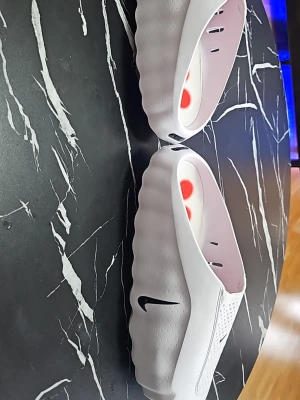 Vita Nike Mind tofflor... - Säljer ett par vita Nike Mind tofflor med svart Swoosh-logga på sidan. Tofflorna har en vågig sula, perforerade detaljer och röda prickar på innersulan. Tillverkade i ett lätt och flexibelt syntetmaterial som ger en modern look.