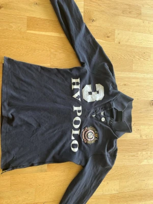 Mörkblå långärmad skjorta HV Polo - Snygg mörkblå långärmad skjorta från HV Polo med stora vita tryck fram och bak, samt broderad logga på bröstet. Skjortan har klassisk krage, knappar vid halsen och sportig stil med siffran 3 på både fram- och baksida. Storleken är s/xs