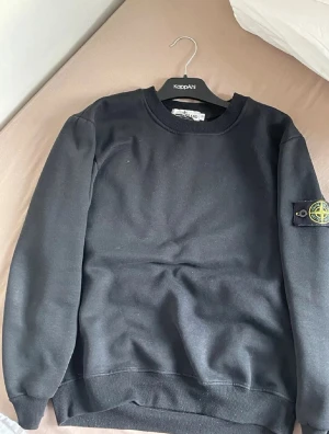 Stone island sweatshirt  - Säljer en svart sweatshirt från Stone Island med klassisk rund hals och ribbade muddar. Tröjan har den ikoniska Stone Island-loggan på vänster ärm. Perfekt för dig som gillar streetwear och enkel stil.