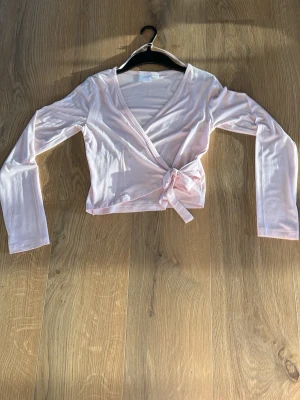 Ljusrosa omlottopp med knyt - Söt ljusrosa omlottopp med långa ärmar och knyt i sidan. Toppen har en v-ringad skärning och är croppad i modellen. Perfekt att styla med höga jeans eller kjol för en trendig look. Nästan aldrig använd.