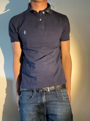 Mörkblå pikétröja från Ralph Lauren - Klassisk mörkblå pikétröja från Ralph Lauren med vit broderad logga på bröstet. Tröjan har korta ärmar, krage och knappar framtill. Tillverkad i mjuk bomull som känns skön mot huden. Perfekt till jeans eller shorts för en avslappnad look.