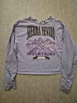 Tröja - Ljuslila långärmad tröja med tryck av berg och texten 'Sierra Nevada Mountains' på bröstet. Tröjan har en avslappnad passform, rund hals med liten v-formad detalj och är tillverkad i mjukt bomullsmaterial. Perfekt för en chill och trendig stil.