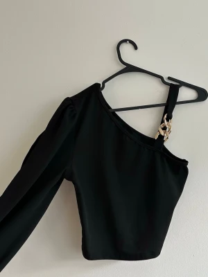 Svart oneshoulder topp med kedjedetalj - Trendig svart oneshoulder topp med lång ärm och snygg kedjedetalj på axelbandet. Toppen har en croppad passform och är tillverkad i ett stretchigt material som sitter snyggt på kroppen. Perfekt för dig som vill sticka ut med en cool detalj.