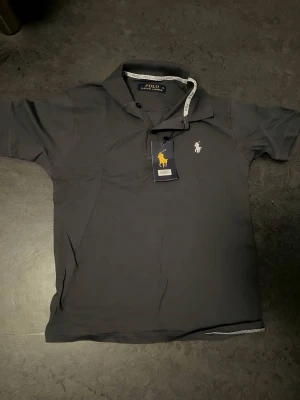 Grå pikétröja från Polo Ralph Lauren - Klassisk svart pikétröja från Polo Ralph Lauren med vit broderad logga på bröstet. Tröjan har krage, korta ärmar och knappar vid halsen. Perfekt till en avslappnad men ändå stilren look. Tröjan har en liten hol som inte syns när man har på. Syns inte utanför tröjan bara inne. Tröjan står m men passar som s-xs ( köpt utomlands så size är annorlunda)
