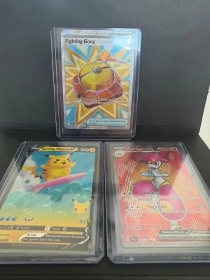 Pokémon spelkort Surfing Pikachu V - Tre Pokémon spelkort i plastfickor: Surfing Pikachu V med blå bakgrund och rosa surfbräda, Fighting Gong med gul och orange motiv, samt Medicham V med röd bakgrund. Perfekt för samlare eller Pokémon-fans som gillar coola och färgglada kort.