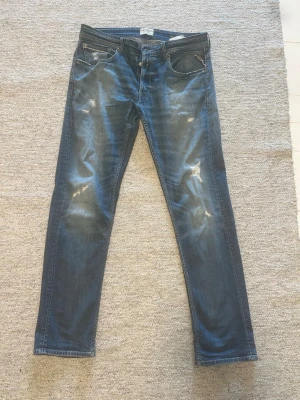 Blå Replay jeans med slitningar - Säljer ett par blå jeans från Replay med snygga slitningar och tvättade detaljer. Klassisk femficksmodell med raka ben och normal midja. Jeansen har en cool, avslappnad look och är tillverkade i bomull med lite stretch för extra komfort.