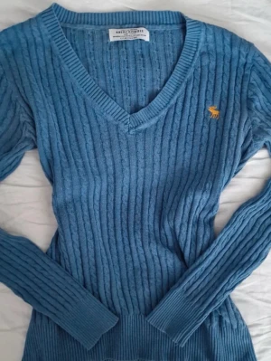 Blå kabelstickad tröja Abercrombie - Snygg blå kabelstickad tröja från Abercrombie & Fitch med V-ringning och gul broderad älg på bröstet. Tröjan har långa ärmar och ribbade muddar. Perfekt för dig som gillar klassisk och stilren stickad design.