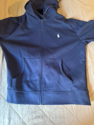 Marinblå ziphoodie från Ralph Lauren - Snygg marinblå hoodie från Ralph Lauren med dragkedja framtill och klassisk vit logga på bröstet. Hoodien har två stora fickor fram och en skön huva med dragsko. Perfekt för en avslappnad och stilren look.