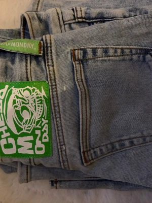 Ljusblåa jeans från Cheap Monday - Säljer ett par ljusblå jeans från Cheap Monday med raka ben och klassisk femficksmodell. Jeansen har en grön patch med grafiskt tryck bak och är tillverkade i denim. Perfekta för dig som gillar streetwear och vill sticka ut med detaljer.