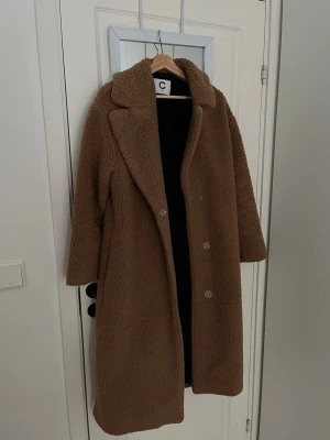 Brun teddyjacka från Cubus - Säljer en lång, brun teddyjacka från C i storlek XS. Jackan har bred krage, knäppning framtill och är gjord i mjukt, fluffigt material som ger en riktigt cozy vibe. Perfekt för kyliga dagar när du vill vara både varm och stylish.