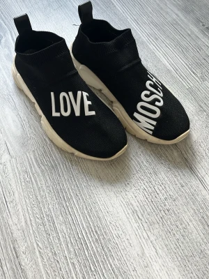 Love Moschino svarta slip-on sneakers - Svarta slip-on sneakers från Love Moschino med vit text på ovandelen. Skorna har en chunky vit sula och är tillverkade i ett stickat material som ger en sock-liknande passform. Perfekta för dig som gillar streetstyle och vill sticka ut.