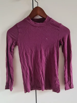 Lila långärmad polotopp Tommy Hilfiger - Lila långärmad polotopp från Tommy Hilfiger med liten logga på bröstet. Tunn och stretchig modell som sitter tight mot kroppen. Perfekt att styla med jeans eller kjol för en trendig look.