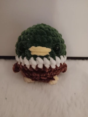 Stickad anka i grönt, vit och brunt garn - Söt handgjord stickad anka med grönt huvud, brunt kropp, vit rand och gula detaljer på näbb och fötter. Mjuk och fluffig, perfekt som dekoration eller present. Svarta plastögon ger ett gulligt uttryck.