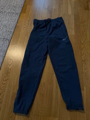 Mörkblå Nike Dri-FIT träningsbyxor - Säljer ett par mörkblå träningsbyxor från Nike med Dri-FIT-material som andas. Byxorna har resår i midjan med dragsko och elastiska benslut. Perfekta för träning eller chill. Diskret Nike-logga på benet.
