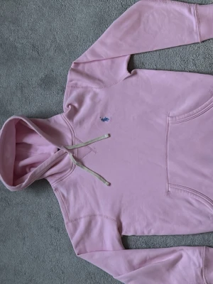 Ljusrosa hoodie från Ralph Lauren - Säljer en ljusrosa hoodie från Ralph Lauren med klassisk huva och dragsnören. Tröjan har en känguruficka framtill och den ikoniska blå loggan broderad på bröstet. Tillverkad i mjukt bomullsmaterial och har ribbade muddar vid ärmar och nederkant. Knappast använd.