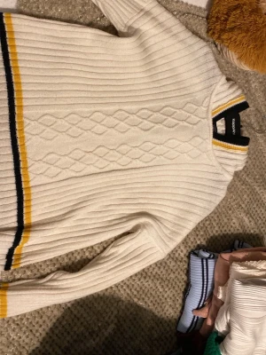 Vit stickad tröja med ränder från Lexington - Klassisk vit stickad tröja från Lexington med ribbad struktur och diskret mönster framtill. Tröjan har gula och marinblå ränder vid krage, ärmslut och nederkant. Långärmad och med en mjuk, bekväm känsla. Perfekt för en preppy look.