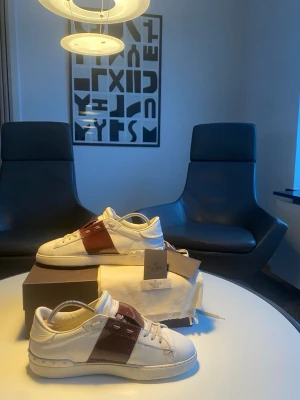 Valentino vita sneakers med vinröd detalj - Snygga vita sneakers från Valentino med en bred, glansig vinröd remsa över sidan och matchande vinröda detaljer vid snörningen. Skorna har klassisk låg profil och är tillverkade i läder med vita snören och sula. Perfekta för dig som vill ha en exklusiv touch på din outfit.