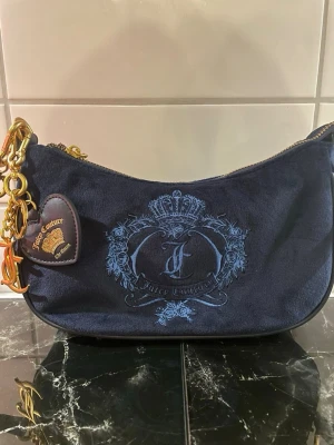 Mörkblå Juicy Couture handväska - Snygg mörkblå handväska från Juicy Couture i mjuk velour med broderad logga och krona framtill. Väskan har guldfärgade detaljer, kedja och ett hjärtformat hänge. Invändigt foder med Juicy Couture-tryck och innerfack med guldbroderad logga.