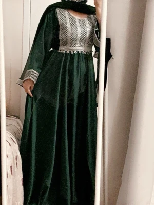 Grön långklänning med silverdetaljer - Elegant grön långklänning med silverfärgat broderi och pärldetaljer vid midjan. Klänningen har långa ärmar med matchande broderi och en flödande siluett. Perfekt för dig som vill sticka ut med en unik och lyxig look.