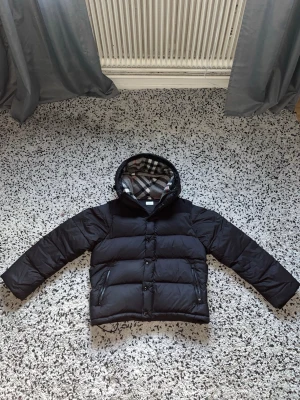 Burberry jacka (S) - Säljer en svart pufferjacka från Burberry med klassiskt rutigt foder i huvan. Jackan har dragkedja och knappar framtill, två fickor med dragkedja och en rejäl huva. Perfekt för kalla vinterdagar och riktigt snygg streetstil.
