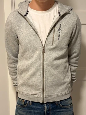 Grå zip-hoodie från Sail Racing - Snygg ljusgrå hoodie med dragkedja från Sail Racing. Tröjan har huva, bröstficka med dragkedja och diskret logga på vänster sida. Tillverkad i mjukt sweatshirtmaterial och har ribbade muddar vid ärmar och midja. Perfekt för en avslappnad stil. Tröjan har några små slitningar på höger bröst dermed priset. Hör av dig vid minsta intresse⭐️