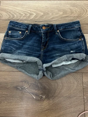 Mörkblå denimshorts med uppvik - Säljer ett par mörkblå denimshorts från Ltb med klassisk femficksdesign och uppvikta benslut. Shortsen har gylf med knapp och dragkedja samt kontrastsömmar. Perfekta för varma dagar och enkel att matcha med olika stilar. Lite stor i storleken