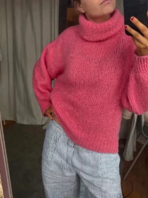 Rosa stickad polotröja - En riktigt snygg rosa stickad polotröja med grov struktur och bred krage. Tröjan har en avslappnad passform och långa ärmar med ribbade muddar. Perfekt för att hålla sig varm och se cool ut under kyliga dagar.