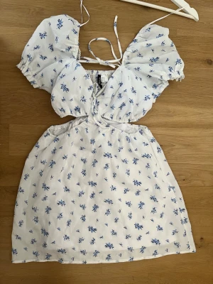 Vit blommig kortklänning med puffärm - Supersöt vit kortklänning med blått blommönster, puffärmar och omlottdetalj framtill. Klänningen har cut-outs i midjan, knytband och smock i ryggen för extra komfort. Perfekt för sommarens alla tillfällen!