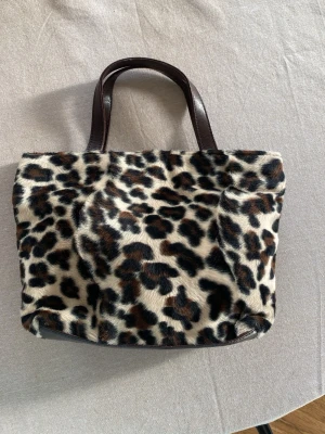 Leopardmönstrad handväska med pälslook - Cool handväska med leopardmönster i brunt, svart och beige. Väskan har korta, mörkbruna handtag och är klädd i ett pälsliknande material som ger en lyxig känsla. Perfekt accessoar för att sticka ut och addera edge till din outfit. Äkta läder!