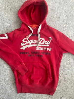 Röd hoodie från Superdry med tryck - Röd hoodie från Superdry med vit logga och japanska tecken på bröstet. Har huva med vita snören, magficka och ribbade muddar. Siffra 7 på ärmen. Mjuk bomullskänsla och sportig streetstil.