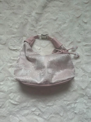 Ljusrosa axelväska med metallicmönster - Snygg ljusrosa handväska i glansigt tyg med silvrigt metallicmönster. Väskan har en kort axelrem i rosa konstläder, metallspänne och dekorativa snören med pärlor. Perfekt för dig som gillar trendiga accessoarer med unik touch.