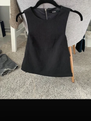 Svart ärmlös topp från Bik Bok - Stilren svart topp från Bik Bok med ärmlös design och rund halsringning. Toppen har en diskret dragkedja i ryggen och är sydd i ett slätt material som ger en snygg passform. Perfekt att matcha med jeans eller kjol för en cool look.