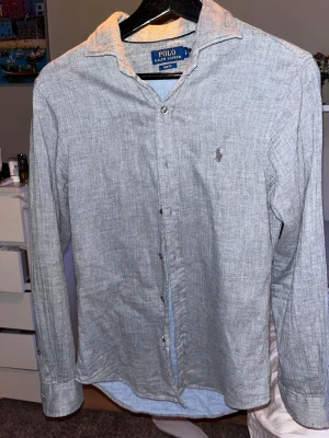 Grå skjorta från Polo Ralph Lauren - Snygg grå skjorta från Polo Ralph Lauren i slim fit-modell. Skjortan har klassisk krage, långa ärmar och diskret broderad logga på bröstet. Tillverkad i ett mjukt och lätt tyg som ger en stilren look.