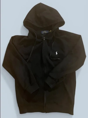 Svart zip up från Polo Ralph Lauren - Säljer en svart hoodie med dragkedja från Polo Ralph Lauren. Klassisk design med huva och dragsnören samt den ikoniska vita logon broderad på bröstet. Perfekt för en avslappnad och stilren look. Det står M men passar bättre som storlek S
