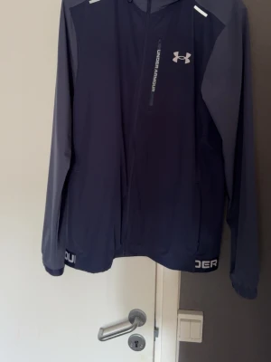 Mörkblå vindjacka från Under Armour - Snygg mörkblå vindjacka från Under Armour med dragkedja framtill och logga på bröstet. Jackan har kontrasterande grå ärmar, bröstficka med dragkedja och vita detaljer vid axlarna. Perfekt för blåsiga dagar och sportiga outfits.