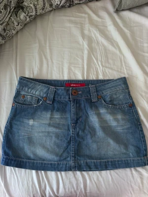 Blå jeanskjol från Gina Tricot - Säljer en klassisk blå jeanskjol från Gina Tricot. Kjolen är kort, har fem fickor, bälteshällor och knappstängning fram. Baksidan har två fickor med rosa broderade detaljer och en liten patch med blommor. Perfekt till sommaren! Storleken säger 30 men passar en M