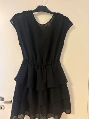 Svart volangklänning med prickigt tyg - Snygg svart klänning med volanger och diskret prickigt tyg. Klänningen har rund halsringning, korta ärmar och markerad midja. Perfekt för dig som gillar en feminin och trendig look.